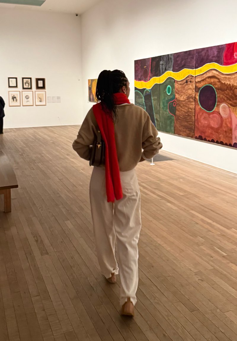 Personne aux cheveux foncés tressés, pantalon beige, pull marron et écharpe rouge marche sur un sol en bois vers des peintures abstraites colorées dans une galerie d'art.