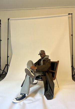 Mann, der einen grauen Hoodie, einen braunen Mantel, eine schwarze Mütze und Sneakers trägt, sitzt auf einem Sessel vor einem weißen Studiohintergrund und passt seine Schuhe an.