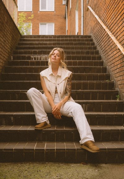 Chaleco de mezclilla beige sin mangas sobre una blusa blanca, combinado con pantalones blancos y zapatos trenzados marrones, sentado en escalones oscuros de azulejos.