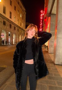 Svart kunstpelsjakke over en tettsittende svart topp, brukt sammen med svarte leggings med nagler. Gateoppsett med et glødende neonskilt i bakgrunnen.