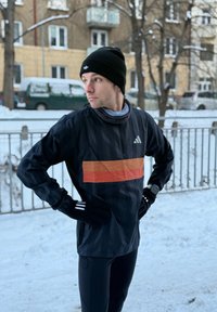 Homme portant un bonnet noir, des gants et une veste sportive avec une bande orange, debout les mains sur les hanches dans un parc urbain enneigé.