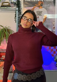 Femme ajustant des lunettes écaille de tortue, portant un pull à col roulé bordeaux et de grandes boucles d'oreilles en anneau, dans une pièce colorée avec des affiches et des plantes.