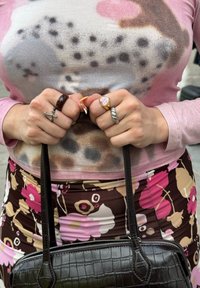 Camisa de manga longa rosa com um padrão de manchas, saia com padrão floral em castanho e rosa, quatro anéis, segurando uma pequena mala preta.