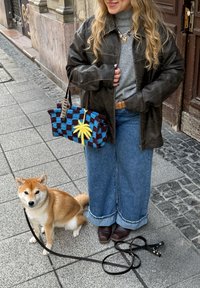 Naisel on seljas nahktagi ja laiade säärtega teksad, seisab tänaval, hoides sinise-pruuni ruudulist kotti, kõrval istub väike beež koer.