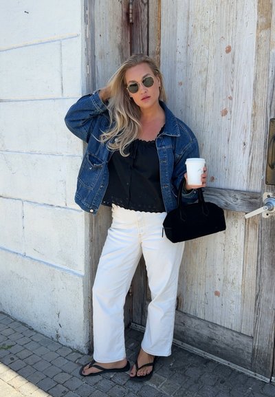 Mujer con cabello rubio largo que lleva chaqueta vaquera, camiseta negra, pantalones blancos y gafas de sol, está junto a una puerta de madera sosteniendo una taza de café y un bolso negro.