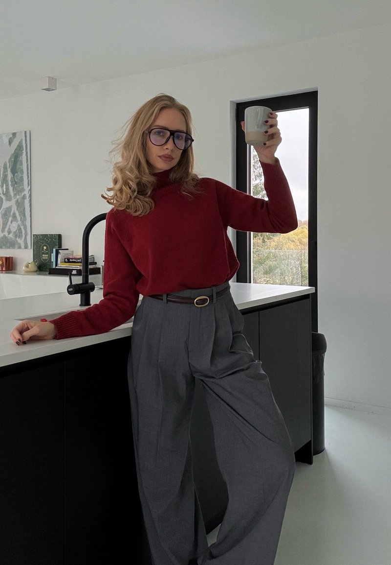 Roter Strickpullover, graue Hosen mit weitem Bein, Glas in der Hand, schwarze Küchenschränke, weiße Arbeitsplatte, große Fenster mit natürlichem Licht.