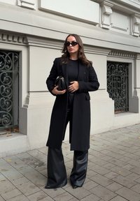 Schwarzer Wollmantel mit taillierter Silhouette, kombiniert mit einem schwarzen, figurbetonten Oberteil und breiten, veganen Lederhosen. Abgerundet mit einer kleinen schwarzen Clutch.