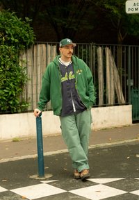 Grønn hettegenser over en mørkeblå baseballskjorte, lysegrønne bukser og brune sko med leopardmønster. Utendørs miljø med planter i bakgrunnen.