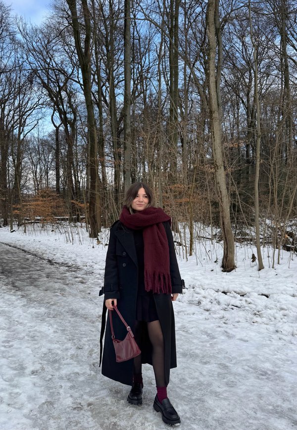 Femme en manteau noir, écharpe bordeaux et sac assorti marchant sur un sentier forestier enneigé avec des arbres nus sous un ciel bleu clair.