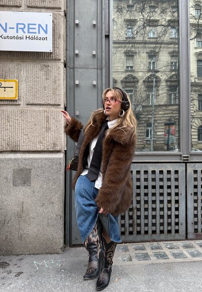 Joven mujer con un abrigo de piel marrón, gafas rojas, corbata negra, auriculares, falda vaquera y botas estampadas de pie en una acera de la ciudad.