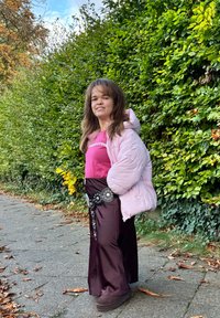 Giacca a vento rosa chiaro, gonna aderente bordeaux con dettagli decorativi in argento, calzature nere, sentiero all'aperto fiancheggiato da vegetazione verde.