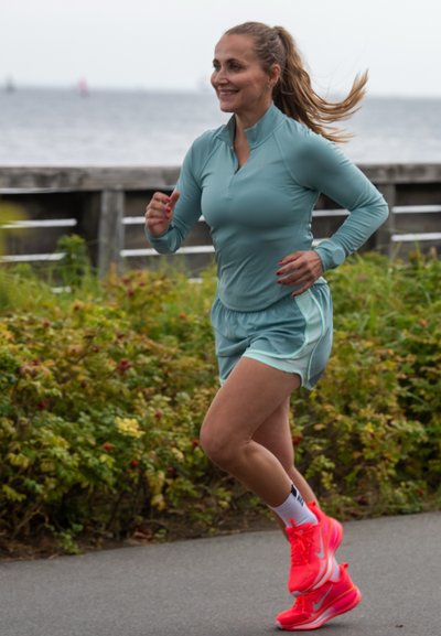 Top deportivo de manga larga azul claro y pantalones cortos a juego. Zapatillas de correr rosa brillante con acentos blancos. Mujer corriendo al aire libre junto al agua.