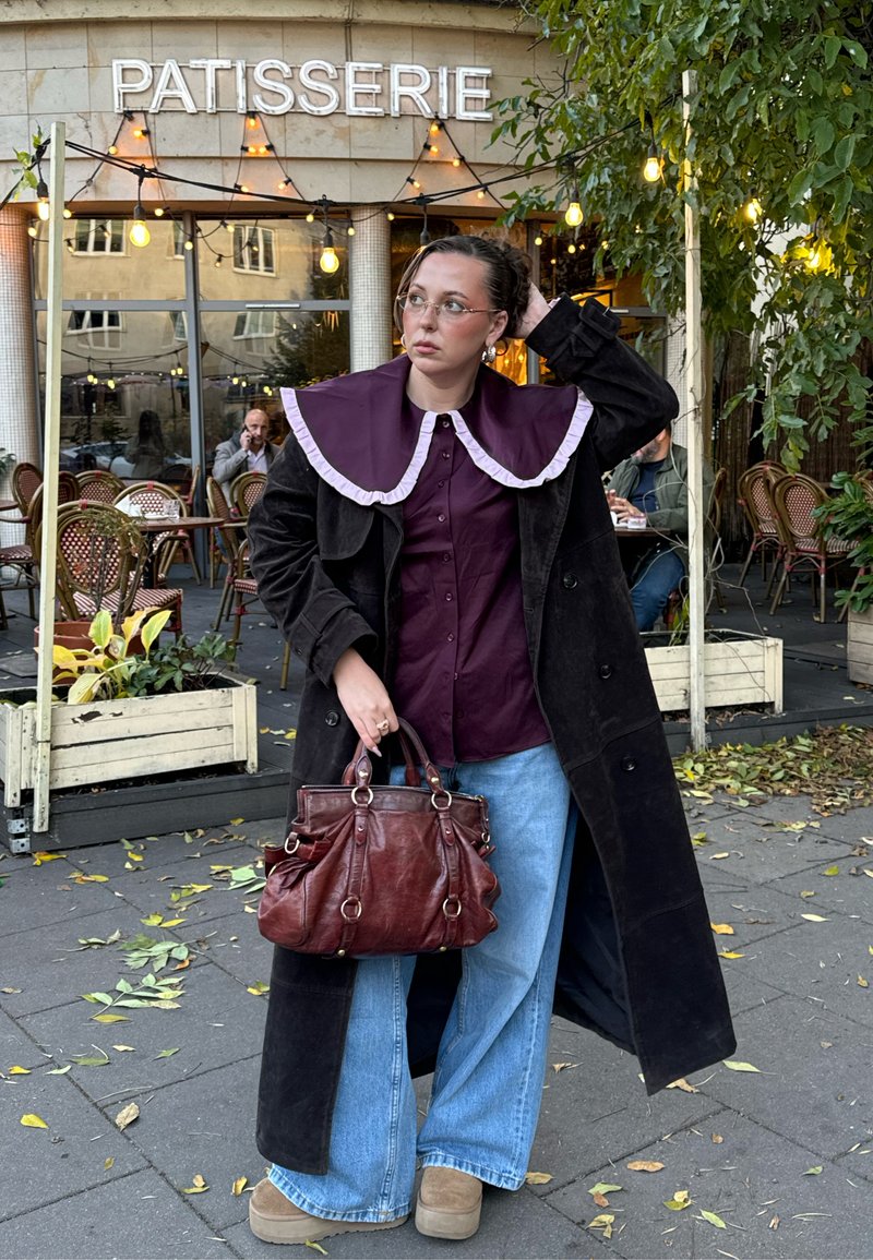 Lungo cappotto nero sopra una camicia color bordeaux con un grande colletto arricciato, jeans larghi blu e una borsa marrone. Ambiente di caffè all'aperto con piante verdi.
