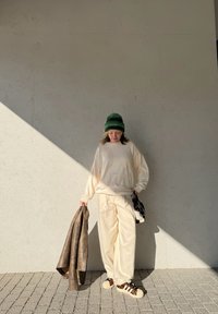 Pulôver oversized bege com padrões texturizados, pantalones bege, casaco castanho pendurado no braço, gorro verde e sapatos estampados sobre pavimento texturizado.