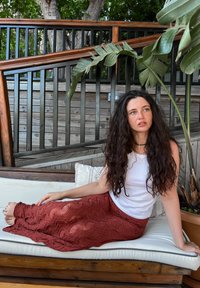 Žena sjedi na svijetlom udobnom kauču, odjevena u bijeli top s naramenicama i dugu suknju od rustikastog, teksturiranog pletenog materijala s uzorkom u valovima.