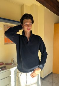 Dark navy knitted sweater with a high collar and ribbed texture, paired with light-coloured trousers. Sunlight accentuates the fabric details.