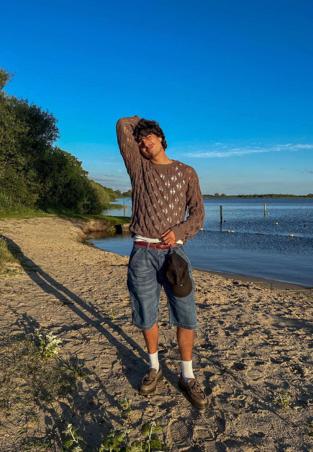 Pullover en tricot marron avec un motif en losange, short en denim bleu, chaussettes blanches et chaussures marron. Debout sur une plage de sable au bord de l'eau, ombre visible.