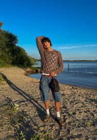 Pullover en tricot marron avec un motif en losange, short en denim bleu, chaussettes blanches et chaussures marron. Debout sur une plage de sable au bord de l'eau, ombre visible.