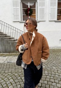 Veste en suède marron avec un col haut et une doublure en shearling, équip ée de grandes poches frontales et de boutons, portée sur une chemise à carreaux et un pantalon sombre.