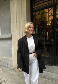 Blazer noir oversized, t-shirt blanc, pantalon blanc taille haute et ceinture noire. Tissu lisse avec une coupe décontractée. Cadre urbain décontracté.