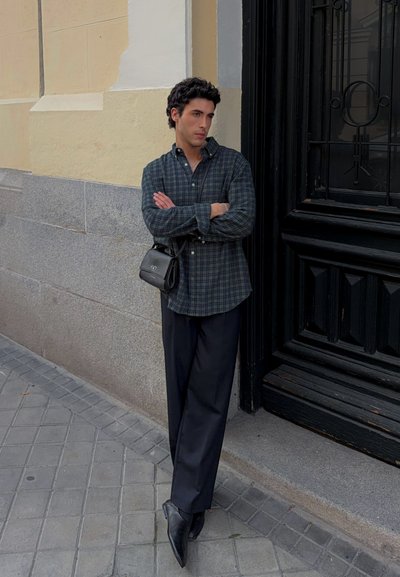 Hombre con camisa de cuadros oscura y pantalones negros se apoya contra una pared beige junto a una puerta negra, con los brazos cruzados y una bolsa negra al hombro.