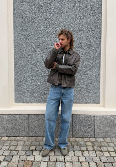 Hombre con cabello rizado que lleva una chaqueta de cuero marrón, jeans azules y zapatos beige, de pie sobre adoquines frente a una pared texturizada gris y crema.