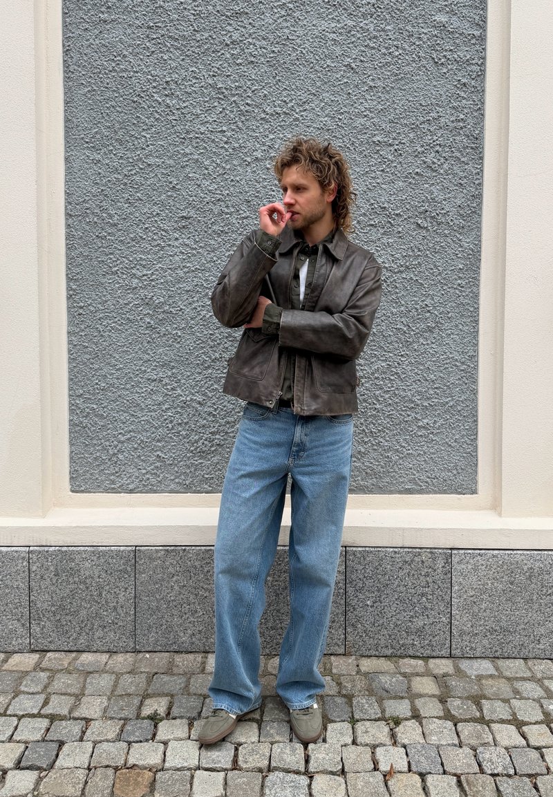 Homme aux cheveux bouclés portant une veste en cuir marron, un jean bleu et des chaussures beige, debout sur des pavés devant un mur texturé gris et crème.