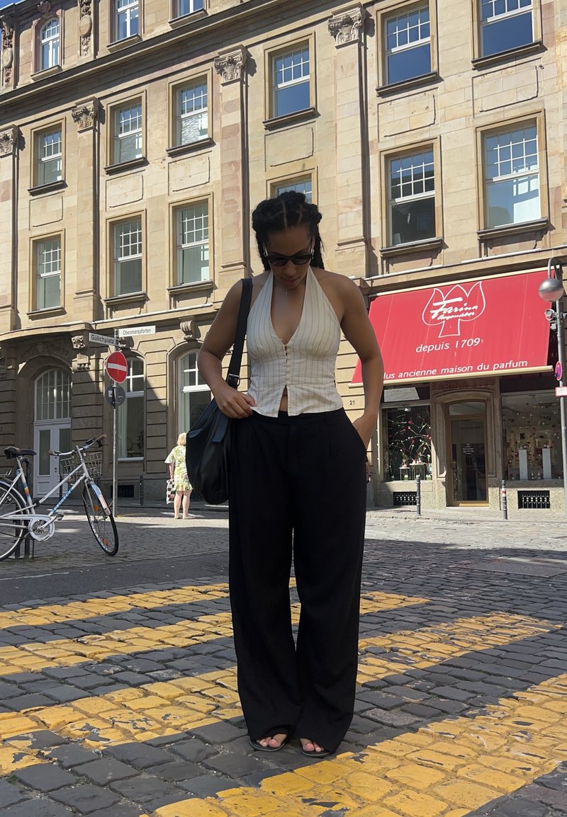 Top a spalla bianco a strisce con bottoni, pantaloni larghi neri e sandali. Sullo sfondo si vedono una strada ciottolata e degli edifici.