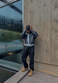 Černá kožená krátká bunda přes bílé tričko, černé džíny a světle hnědé pracovní boty. V pozadí je texturovaná skleněná stěna připomínající vodu.