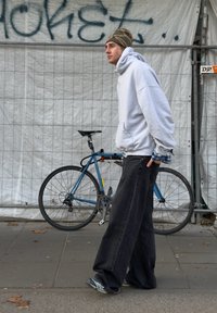 Sweat-shirt gris à capuche, pantalons noirs amples, chaussures de sport. En arrière-plan, un vélo bleu et un mur blanc couvert de graffitis.