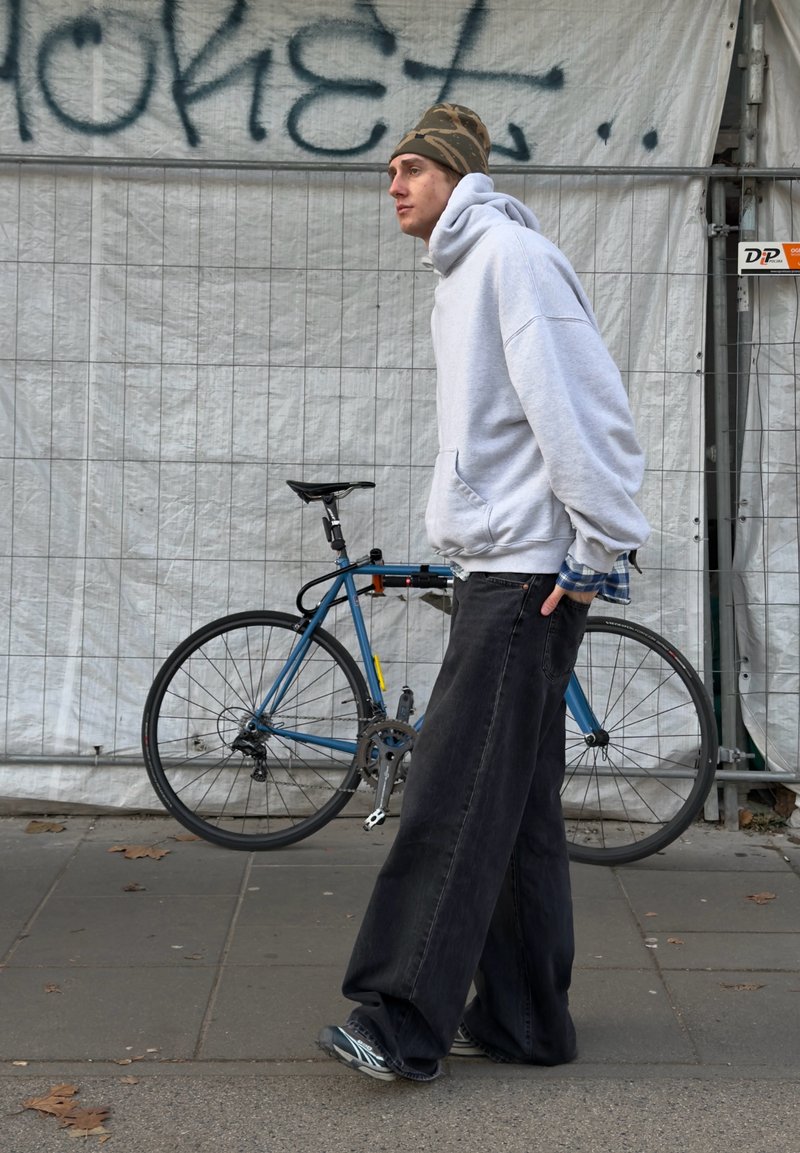 Sweat-shirt gris à capuche, pantalons noirs amples, chaussures de sport. En arrière-plan, un vélo bleu et un mur blanc couvert de graffitis.