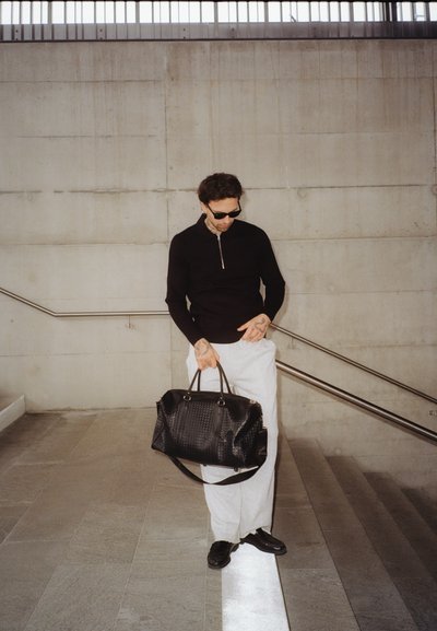 Sudadera negra con cremallera, pantalones gris claro, bolso de cuero negro elegante con patrón texturizado y zapatos oscuros. Fondo de hormigón liso.