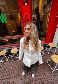 Veste blanche portée sur un pantalon marron ample, avec des cheveux bouclés. En arrière-plan, des encadrements de fenêtres rouges et des tabourets en bois sur un pavement en briques.