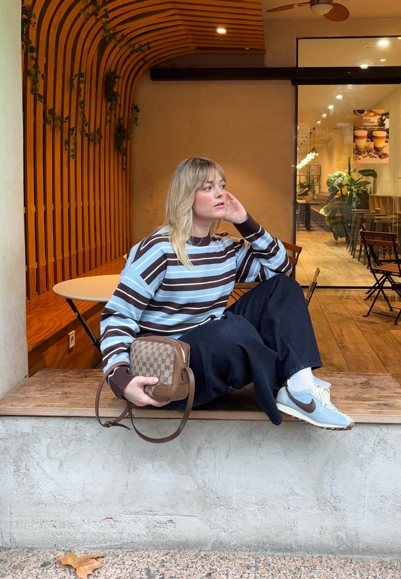 Femme portant un pull rayé et un pantalon foncé, assise sur un rebord en bois à l'extérieur d'un café, tenant un petit sac à main marron, regardant sur le côté.