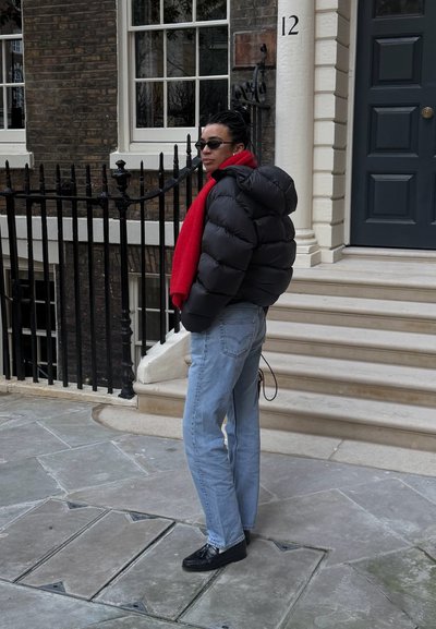 Persona con chaqueta acolchada negra y bufanda roja de pie en la acera cerca de una barandilla negra y escalones que conducen a un edificio con puerta azul marcado con el número 12.