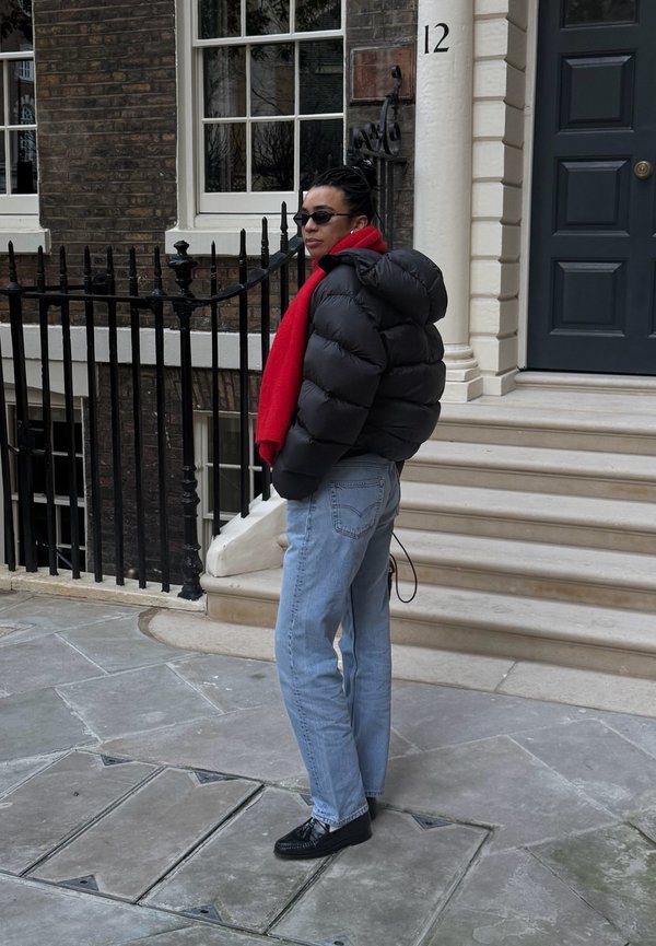 Osoba v čiernej prešívanej bunde a červenom šále stojí na chodníku pri čiernom zábradlí a schodoch vedúcich k budove s modrými dverami a číslom 12.