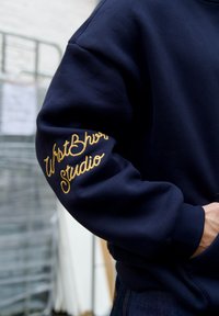 Sweat-shirt bleu marine avec texte brodé en or "WristWatch Studio" sur la manche gauche. Tissu épais avec une coupe décontractée.