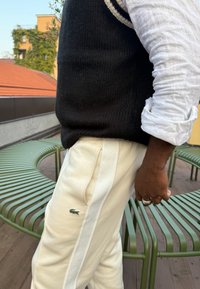 Gilet nero lavorato a maglia su una camicia bianca in lino, abbinato a pantaloni color crema con un logo verde e strisce bianche sui lati.