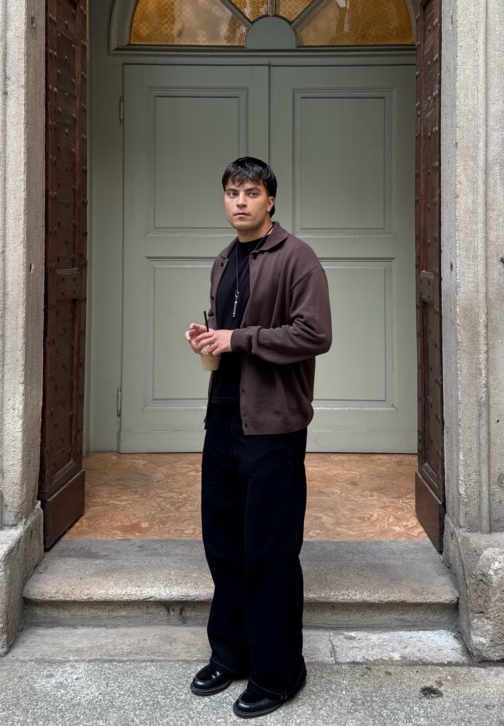 Hnědý svetr přes černé tričko, černé široké kalhoty a černé boty. Drží nápoj, stojí na kamených schodech u dvoří s dvojitými dveřmi.