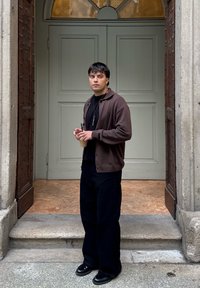 Hnědý svetr přes černé tričko, černé široké kalhoty a černé boty. Drží nápoj, stojí na kamených schodech u dvoří s dvojitými dveřmi.