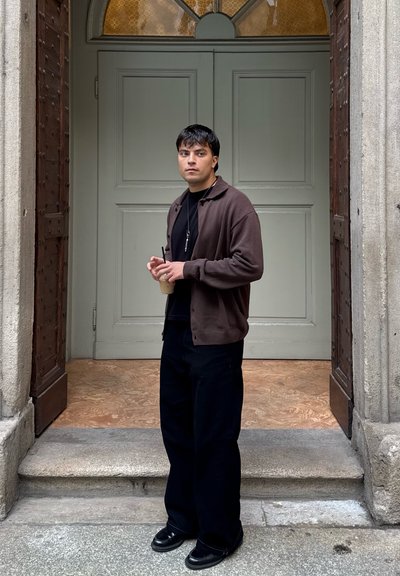 Cardigan marrón sobre una camiseta negra, pantalones anchos negros y zapatos negros. Sosteniendo una bebida, de pie en escalones de piedra junto a una puerta doble.