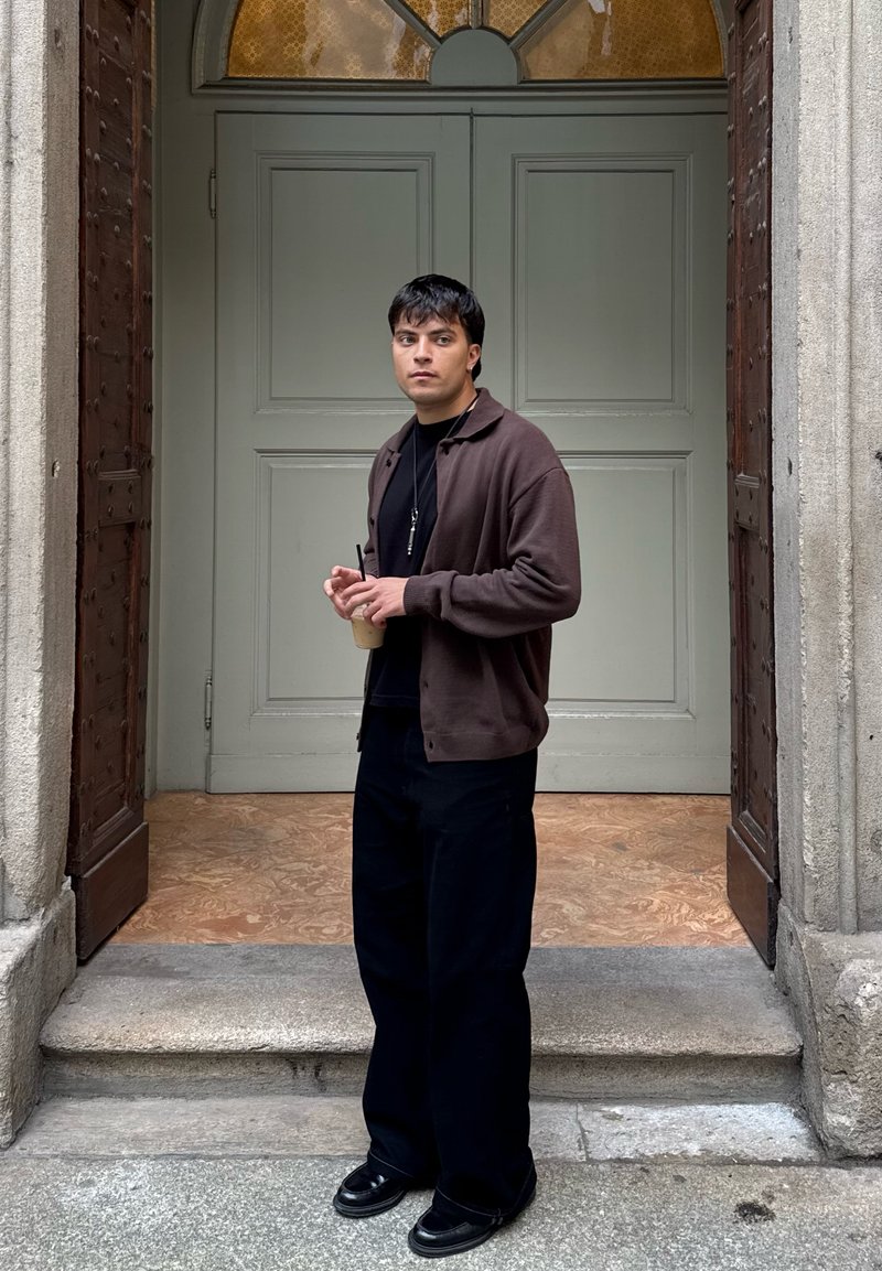 Cardigan marrone sopra una maglietta nera, pantaloni neri a gamba larga e scarpe nere. Tiene una bevanda, in piedi su scalini di pietra accanto a una porta doppia.