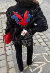 Jaqueta de denim preta com um grande logótipo da Playboy em vermelho e azul nas costas, combinada com jeans pretos boca de sino e uma pequena bolsa vermelha.