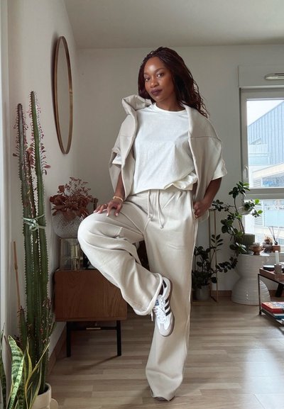 Mujer con pantalones deportivos beige y zapatillas blancas, de pie sobre una pierna en una habitación luminosa con plantas, un armario de madera y un espejo de pared redondo.