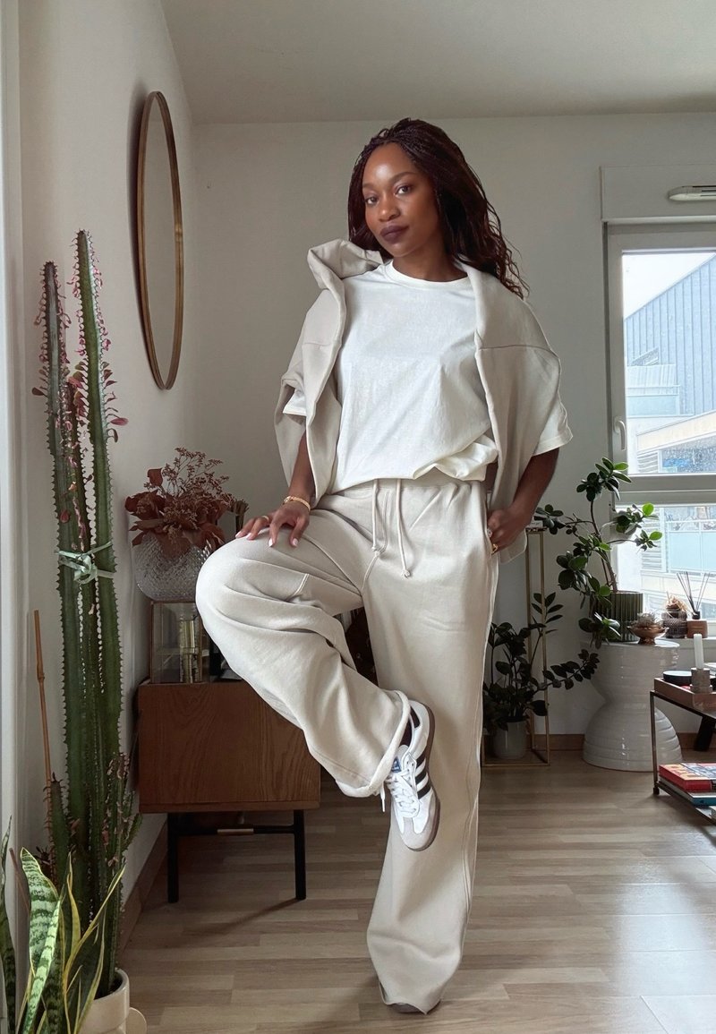 Žena u bež trenirci i bijelim tenisicama stoji na jednoj nozi u svijetloj sobi s biljkama, drvenim ormarom i okruglim zidnim ogledalom.