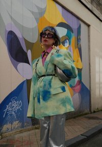 Υπερμεγέθης, παστέλ μπλε-πράσινη ζακέτα με εφέ tie-dye, συνδυασμένη με γυαλιστερές ασημένιες παντελόνες σε φαρδιά γραμμή και μεγάλα σκουλαρίκια σε σχήμα αστεριού.