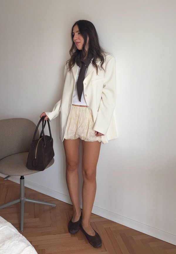 Vit blazer över en vit topp, beige strukturerade shorts och mörkbruna skor. Brun handväska med guldiga detaljer. Minimalistisk stil och neutrala färger.