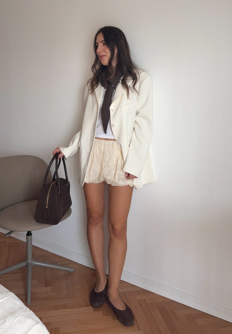 Weißer Blazer über einem weißen Oberteil, beigen strukturierten Shorts und dunkelbraunen Schuhen. Braune Handtasche mit goldenen Beschlägen. Minimalistischer Stil und neutrale Farben.