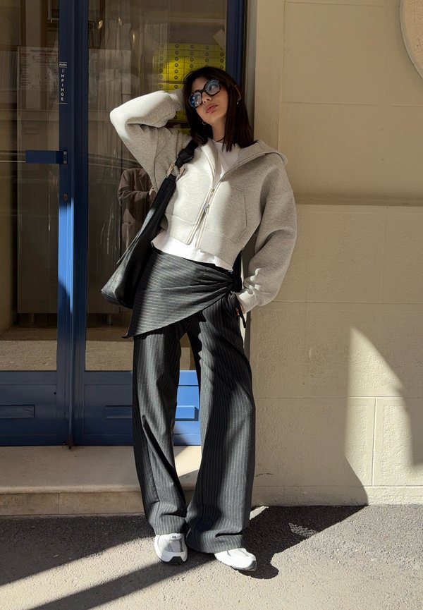 Kvinne iført solbriller, lys grå hettejakke med glidelås, svarte pinstripe-bukser med skjørtedetalj, hvite joggesko og svart skulderveske, stående ved en blå dør.
