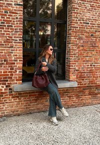 Κουβερτισμένη μπορντό τσάντα χεριού, γκρι σακάκι πάνω από μαύρη μπλούζα, πράσινα καμπάνα παντελόνια με άσπρες τελείες, γκρι αθλητικά παπούτσια και φόντο από τούβλινο τοίχο.